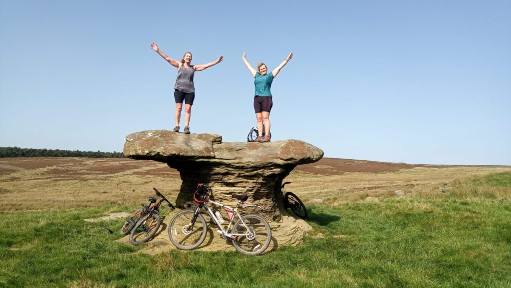 Two women standing on a big stone with bikes underneath.