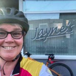 Jayne 1 smiling woman in bike helmet in front of window with a sign saying Jayne's.