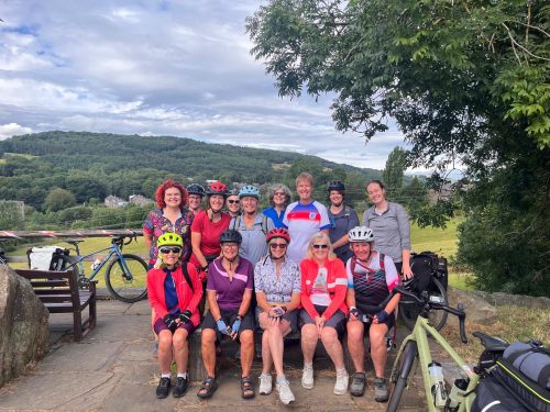 A group of thirteen women, most wearing cycling gear and helmets, pose together outdoors with bikes visible in the background. Some are sitting on a bench while others stand behind them, all smiling. The setting is a scenic countryside with green hills, trees, and a cloudy sky.